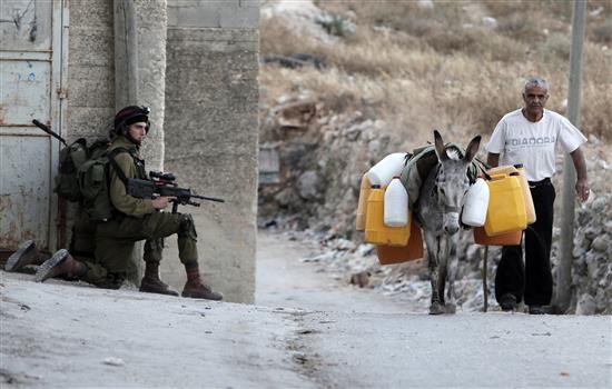 فلسطيني ينقل المياه فيما يتمركز جنديان إسرائيليان خلف حائط في قرية عورتا في الضفة الغربية أمس (أ ف ب)